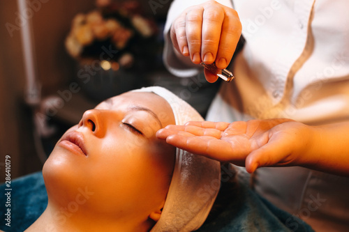 Hands of a cosmetologist pouring from a ampoule skin care product which will apply on a female face.