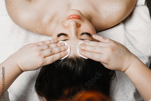 Upper view of a cosmetologist hand doing skin care routine before facial treatment on a cute female face in wellness center.