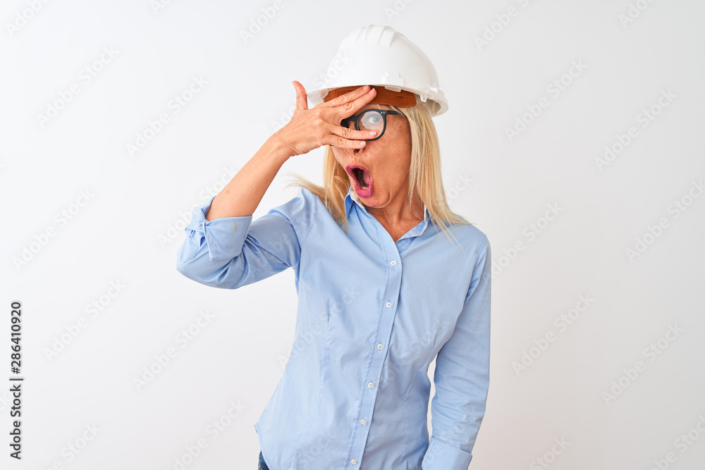 Middle age architect woman wearing glasses and helmet over isolated white background peeking in shock covering face and eyes with hand, looking through fingers with embarrassed expression. Middle age architect woman wearing glasses and helmet over isolated white background peeking in shock covering face and eyes with hand, looking through fingers with embarrassed expression.