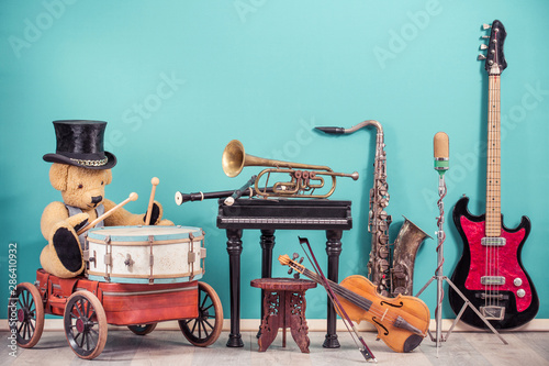 Canvas Print Retro Teddy Bear toy in old cylinder hat sitting on old luggage with wheels and playing the drum and musical instruments set front mint blue wall background