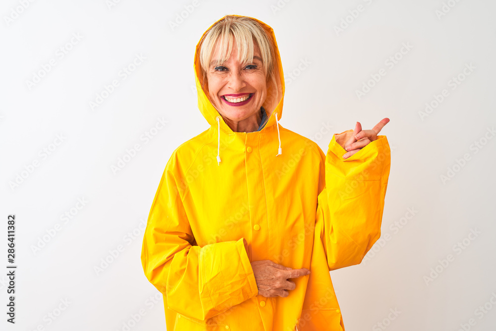 Middle age woman wearing rain coat with hood standing over isolated white background with a big smile on face, pointing with hand and finger to the side looking at the camera. Middle age woman wearing rain coat with hood standing over isolated white background with a big smile on face, pointing with hand and finger to the side looking at the camera.