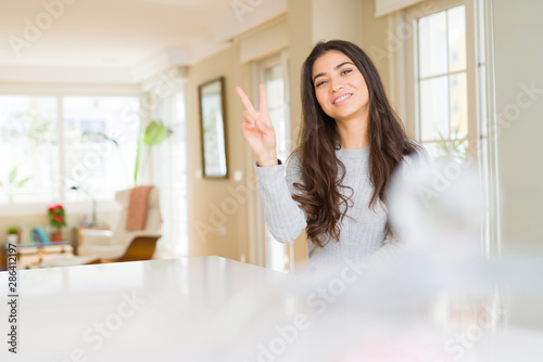 Young beautiful woman at home smiling with happy face winking at the camera doing victory sign. Number two.