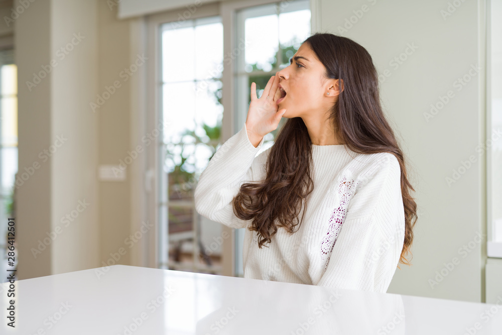 Young beautiful woman at home on white table shouting and screaming loud to side with hand on mouth. Communication concept. Young beautiful woman at home on white table shouting and screaming loud to side with hand on mouth. Communication concept.
