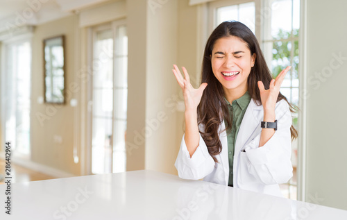 Young woman wearing medical coat at the clinic as therapist or doctor celebrating mad and crazy for success with arms raised and closed eyes screaming excited. Winner concept Free Download Young woman wearing medical coat at the clinic as therapist or doctor celebrating mad and crazy for success with arms raised and closed eyes screaming excited. Winner concept - GFXLoad