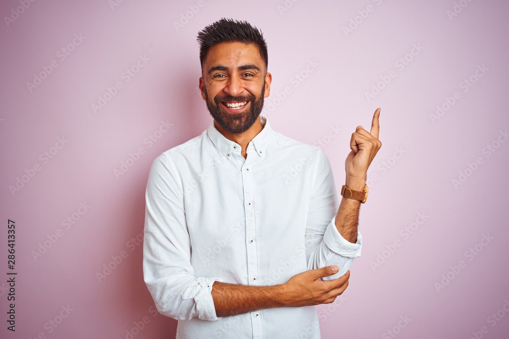 Young indian businessman wearing elegant shirt standing over isolated pink background with a big smile on face, pointing with hand and finger to the side looking at the camera.
