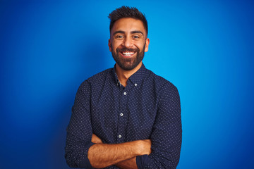 Young indian elegant man wearing shirt standing over isolated blue background happy face smiling with crossed arms looking at the camera. Positive person.