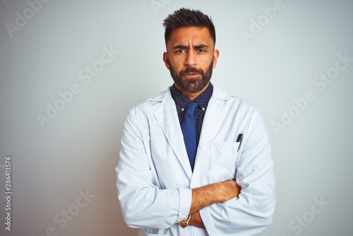 Young indian doctor man standing over isolated white background skeptic and nervous, disapproving expression on face with crossed arms. Negative person.