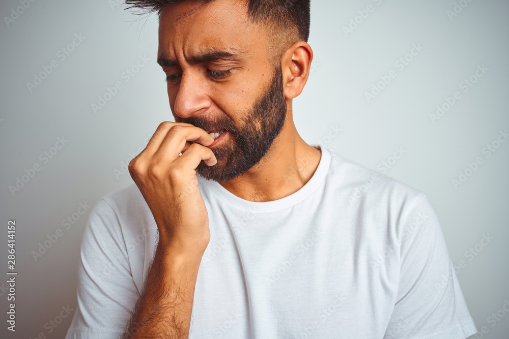 Young indian man wearing t-shirt standing over isolated white background looking stressed and nervous with hands on mouth biting nails. Anxiety problem. Young indian man wearing t-shirt standing over isolated white background looking stressed and nervous with hands on mouth biting nails. Anxiety problem.