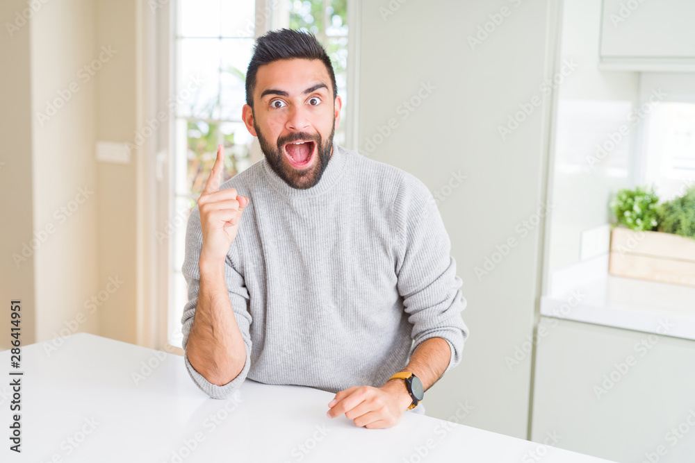 Handsome hispanic man wearing casual sweater at home pointing finger up with successful idea. Exited and happy. Number one.