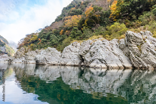 Pale gray rocks and green, yellow and orange trees are reflected in the deep green of the Yoshino river in Japan © Chris Anderson 