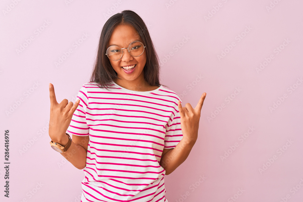 Young chinese woman wearing striped t-shirt and glasses over isolated pink background shouting with crazy expression doing rock symbol with hands up. Music star. Heavy concept. Young chinese woman wearing striped t-shirt and glasses over isolated pink background shouting with crazy expression doing rock symbol with hands up. Music star. Heavy concept.