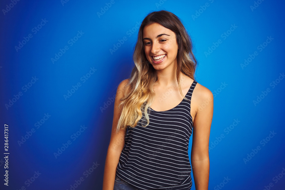 Young beautiful woman wearing stripes t-shirt over blue isolated background looking away to side with smile on face, natural expression. Laughing confident. Young beautiful woman wearing stripes t-shirt over blue isolated background looking away to side with smile on face, natural expression. Laughing confident.