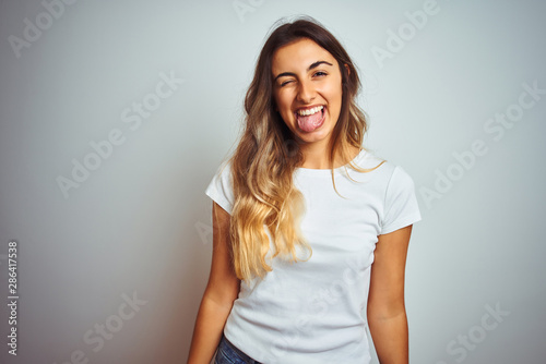 Young beautiful woman wearing casual white t-shirt over isolated background sticking tongue out happy with funny expression. Emotion concept.