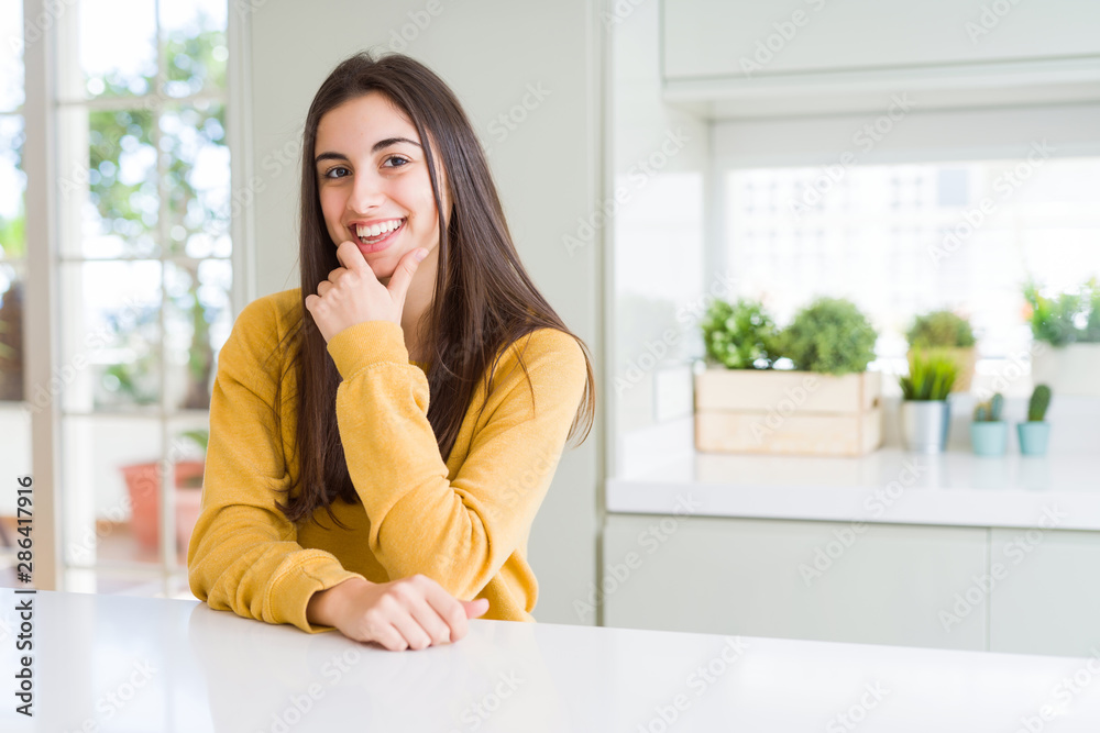 Beautiful young woman wearing yellow sweater looking confident at the camera with smile with crossed arms and hand raised on chin. Thinking positive.