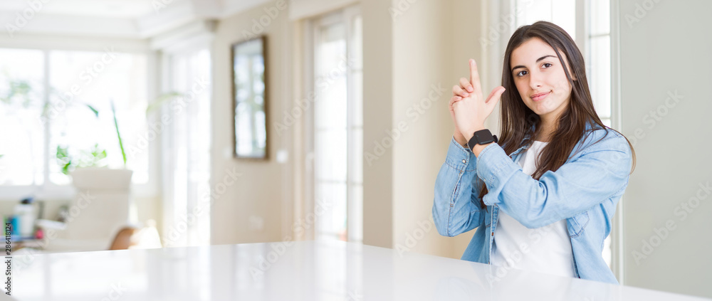 Wide angle picture of beautiful young woman sitting on white table at home Holding symbolic gun with hand gesture, playing killing shooting weapons, angry face