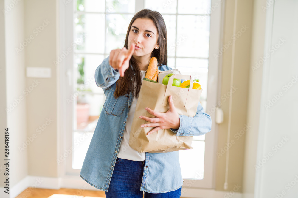 Beautiful young woman holding paper bag full of healthy groceries pointing with finger to the camera and to you, hand sign, positive and confident gesture from the front Beautiful young woman holding paper bag full of healthy groceries pointing with finger to the camera and to you, hand sign, positive and confident gesture from the front