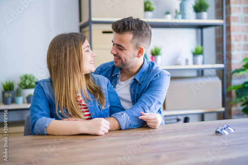 Young beautiful couple sitting on the table at home, hugging in love very happy for moving to new home with cardboard boxes behind them