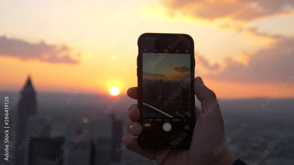 Person photographing/filming sunset in city with a smartphone. Tourist taking photo of sundown.