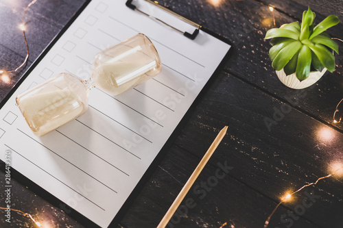 Foto clipboard with blank list with cases to tick off on wooden desk with hourglass