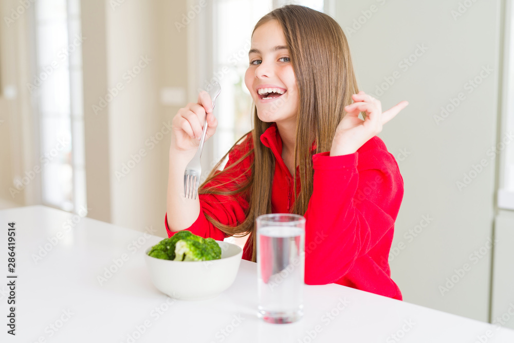 Beautiful young girl kid eating fresh broccoli and drinking water very happy pointing with hand and finger to the side Beautiful young girl kid eating fresh broccoli and drinking water very happy pointing with hand and finger to the side