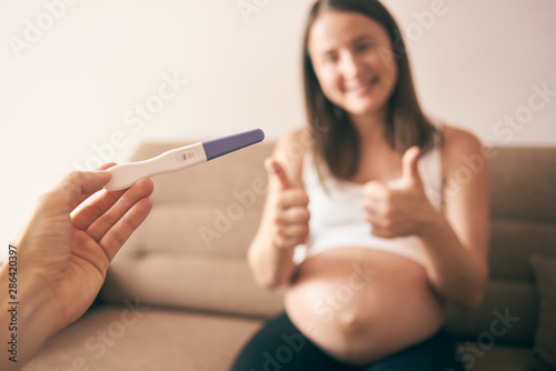 Selective focus of pregnancy test in male hands and cheerful pregnant woman at background. Smiling future mother shoring sign ok with hands and expecting little baby. Concept of happiness.