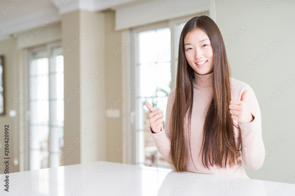 Beautiful Asian woman wearing casual sweater on white table success sign doing positive gesture with hand, thumbs up smiling and happy. Looking at the camera with cheerful expression, winner gesture. Beautiful Asian woman wearing casual sweater on white table success sign doing positive gesture with hand, thumbs up smiling and happy. Looking at the camera with cheerful expression, winner gesture.