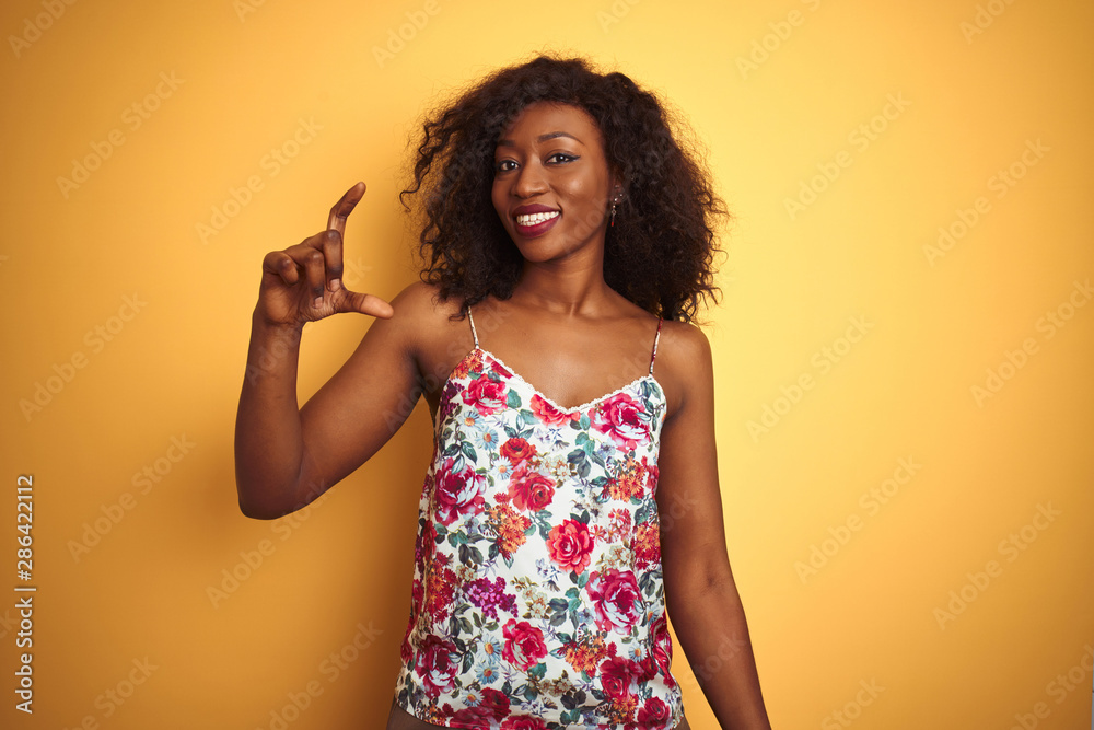 African american woman wearing floral summer t-shirt over isolated yellow background smiling and confident gesturing with hand doing small size sign with fingers looking and the camera. African american woman wearing floral summer t-shirt over isolated yellow background smiling and confident gesturing with hand doing small size sign with fingers looking and the camera.