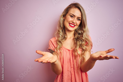 Young beautiful woman wearing t-shirt standing over pink isolated background smiling cheerful offering hands giving assistance and acceptance.