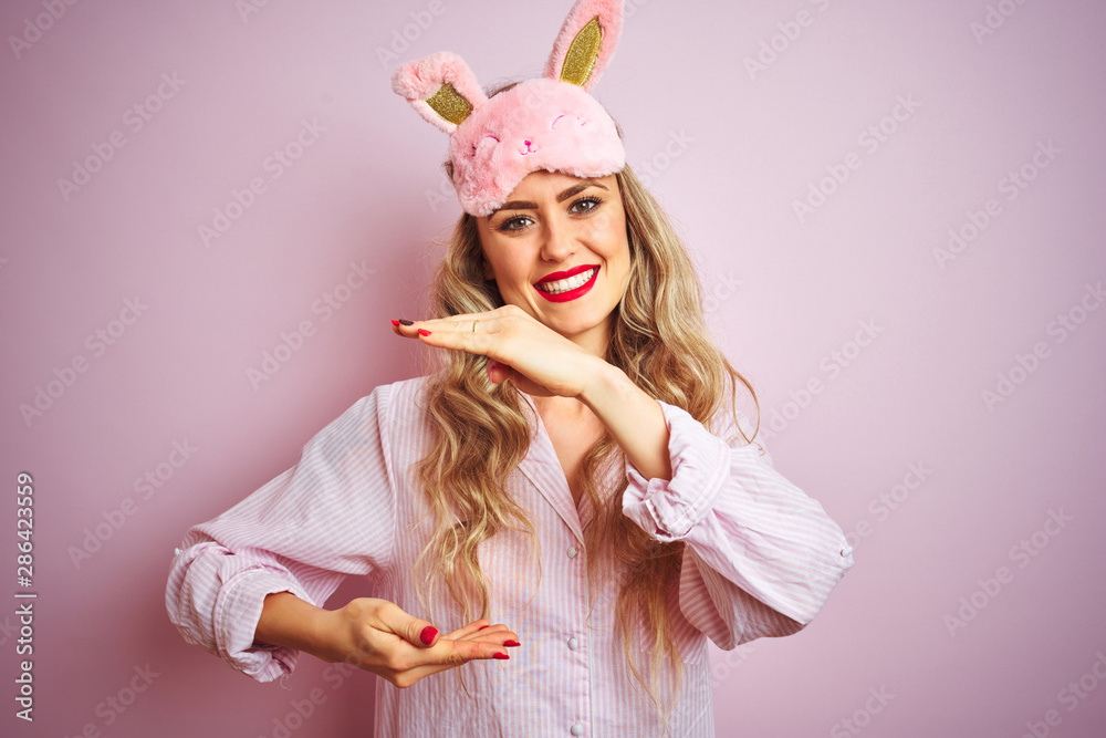 Young beautiful woman wearing pajama and sleep mask over pink isolated background gesturing with hands showing big and large size sign, measure symbol. Smiling looking at the camera. 