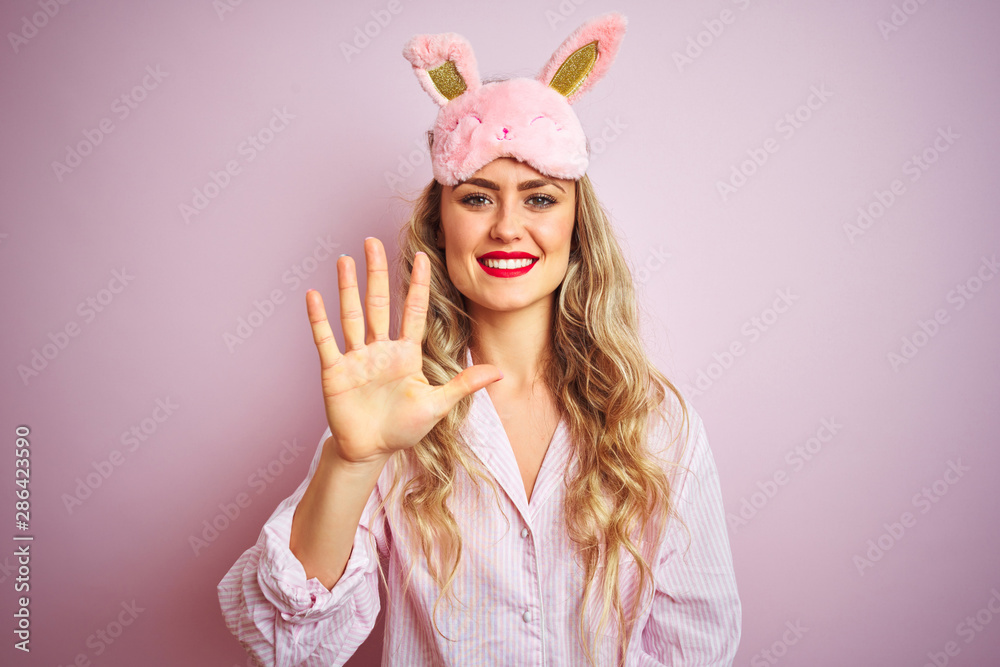 Young beautiful woman wearing pajama and sleep mask over pink isolated background showing and pointing up with fingers number five while smiling confident and happy.