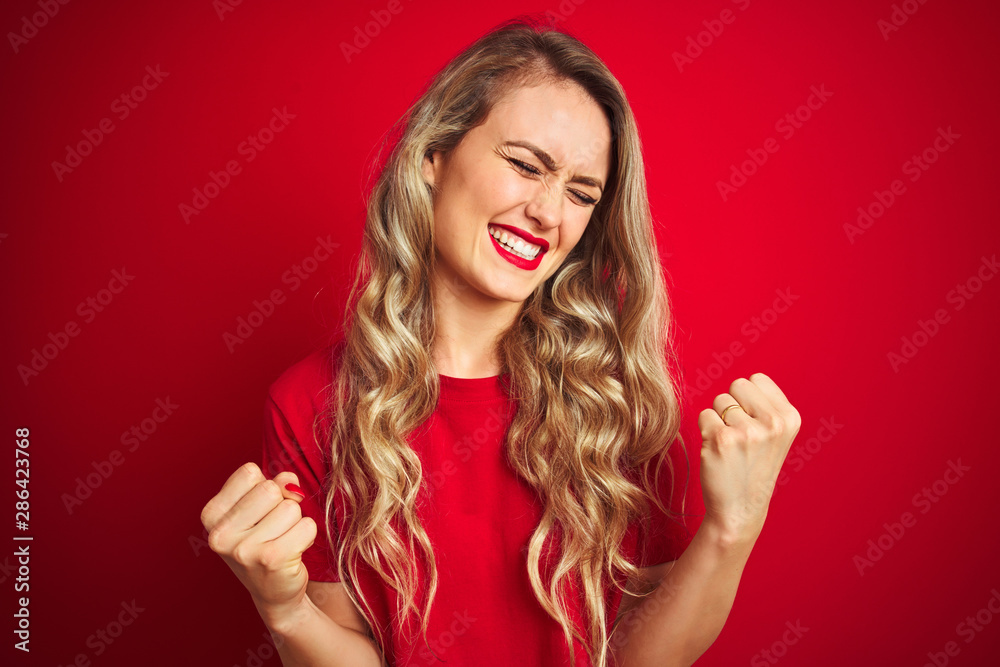 Young beautiful woman wearing basic t-shirt standing over red isolated background very happy and excited doing winner gesture with arms raised, smiling and screaming for success. Celebration concept. Young beautiful woman wearing basic t-shirt standing over red isolated background very happy and excited doing winner gesture with arms raised, smiling and screaming for success. Celebration concept.