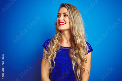 Young beautiful woman standing over blue isolated background looking away to side with smile on face, natural expression. Laughing confident.