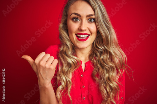 Young beautiful woman standing over red isolated background pointing and showing with thumb up to the side with happy face smiling