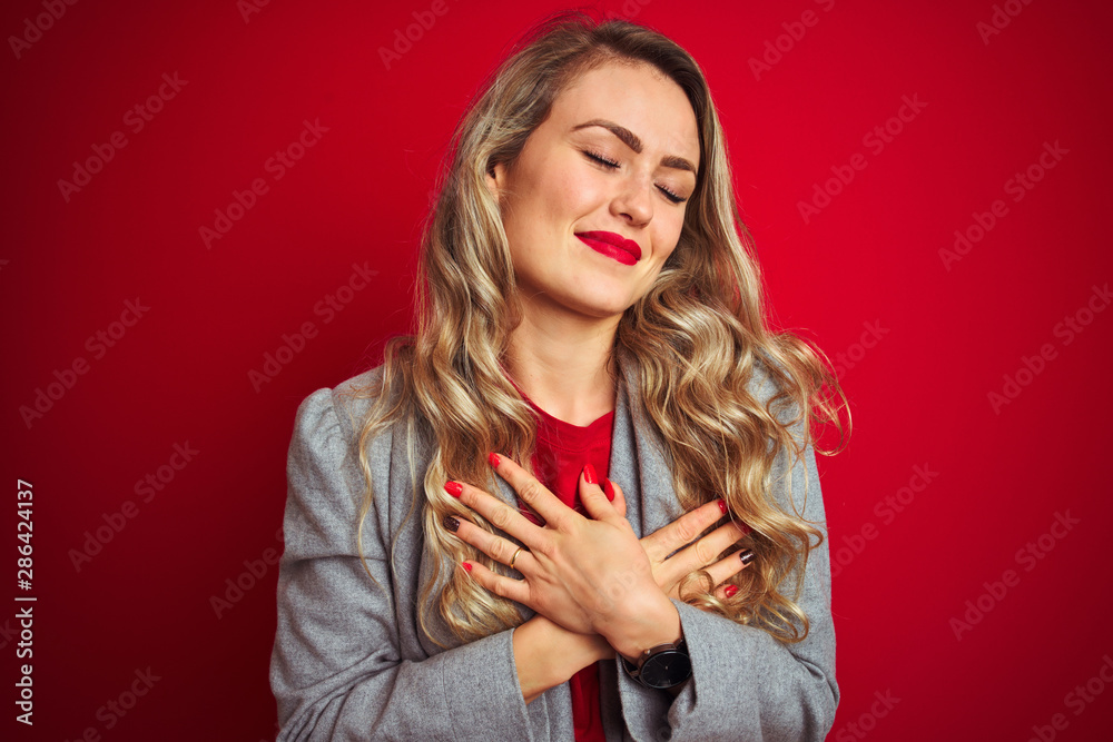 Young beautiful business woman wearing elegant jacket standing over red isolated background smiling with hands on chest with closed eyes and grateful gesture on face. Health concept.
