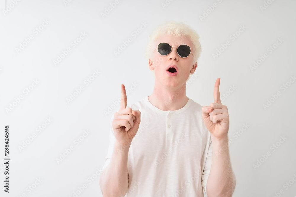 Young albino blond man wearing t-shirt and sunglasses over isolated white background amazed and surprised looking up and pointing with fingers and raised arms. Young albino blond man wearing t-shirt and sunglasses over isolated white background amazed and surprised looking up and pointing with fingers and raised arms.