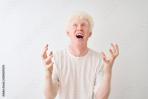 Young albino blond man wearing casual t-shirt standing over isolated white background crazy and mad shouting and yelling with aggressive expression and arms raised. Frustration concept.