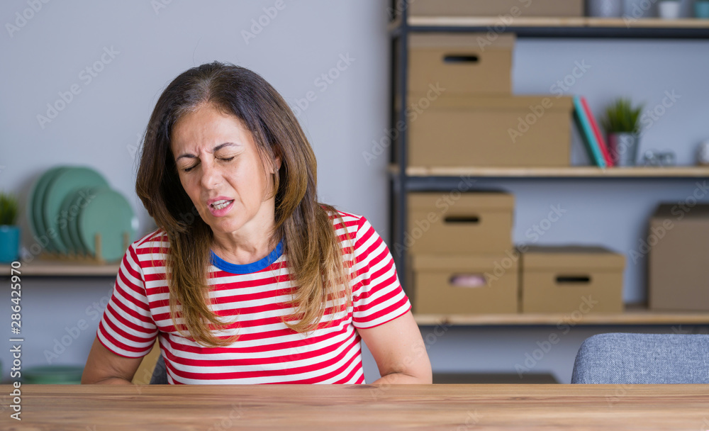 Middle age senior woman sitting at the table at home with hand on stomach because indigestion, painful illness feeling unwell. Ache concept. Middle age senior woman sitting at the table at home with hand on stomach because indigestion, painful illness feeling unwell. Ache concept.