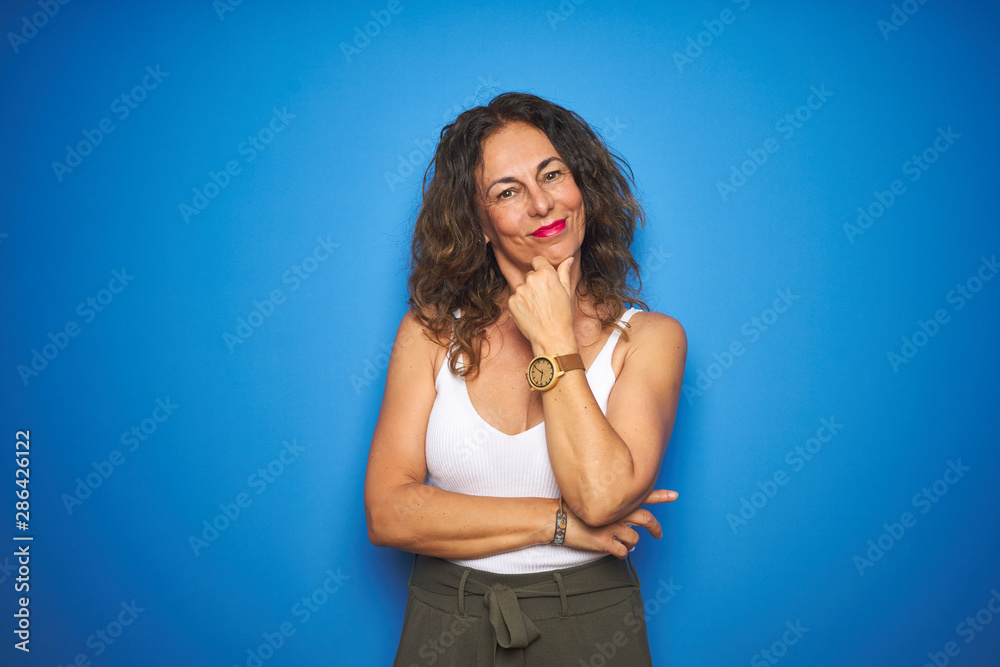 Middle age senior woman with curly hair standing over blue isolated background looking confident at the camera smiling with crossed arms and hand raised on chin. Thinking positive. Middle age senior woman with curly hair standing over blue isolated background looking confident at the camera smiling with crossed arms and hand raised on chin. Thinking positive.