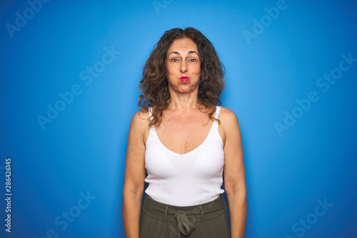 Middle age senior woman with curly hair standing over blue isolated background puffing cheeks with funny face. Mouth inflated with air, crazy expression.