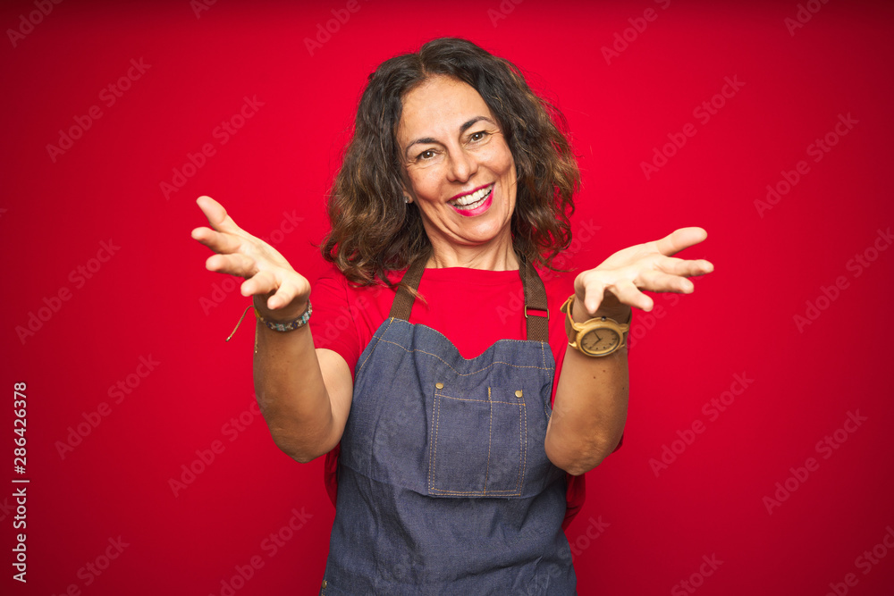 Middle age senior woman wearing apron uniform over red isolated background smiling cheerful offering hands giving assistance and acceptance. Middle age senior woman wearing apron uniform over red isolated background smiling cheerful offering hands giving assistance and acceptance.