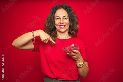 Middle age senior woman eating raspberries over red isolated background with surprise face pointing finger to himself