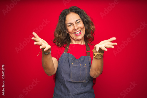 Middle age senior woman wearing apron uniform over red isolated background smiling cheerful offering hands giving assistance and acceptance.