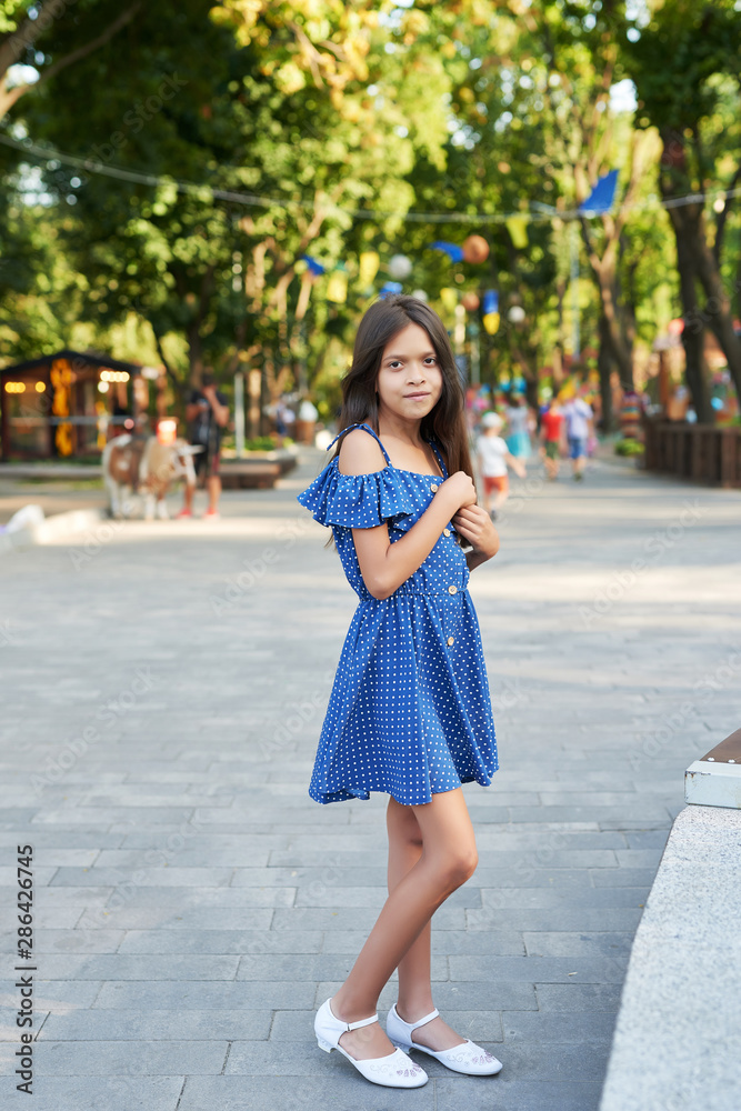 teenager girl in a blue dress walks in the summer park at sunset