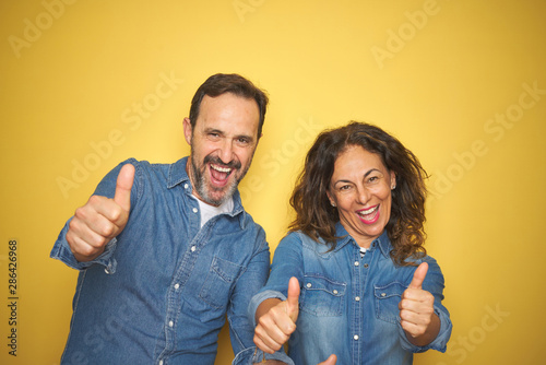 Beautiful middle age couple together standing over isolated yellow background approving doing positive gesture with hand, thumbs up smiling and happy for success. Winner gesture.