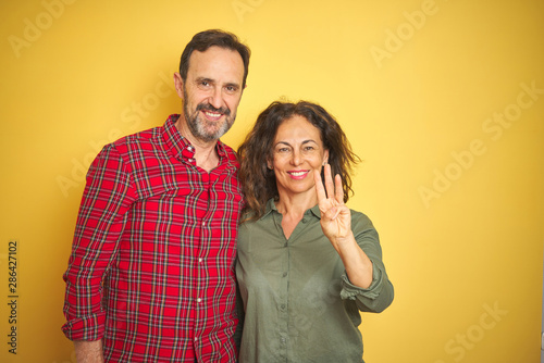 Beautiful middle age couple over isolated yellow background showing and pointing up with fingers number three while smiling confident and happy.