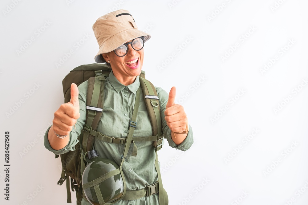 Fototapeta premium Middle age hiker woman wearing backpack canteen hat glasses over isolated white background approving doing positive gesture with hand, thumbs up smiling and happy for success. Winner gesture.