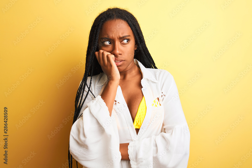 African american woman on vacation wearing bikini and shirt over isolated yellow background looking stressed and nervous with hands on mouth biting nails. Anxiety problem. African american woman on vacation wearing bikini and shirt over isolated yellow background looking stressed and nervous with hands on mouth biting nails. Anxiety problem.