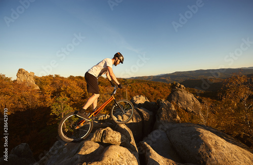 Professional cyclist balancing on trial bicycle on top of big rock, male biker making acrobatic stunt on summer evening, blue sky and forest on background. Concept of extreme sport active lifestyle