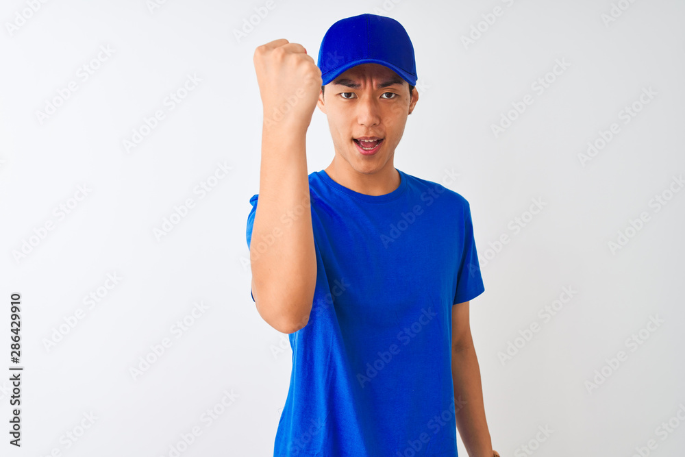 Chinese deliveryman wearing blue t-shirt and cap standing over isolated white background angry and mad raising fist frustrated and furious while shouting with anger. Rage and aggressive concept.