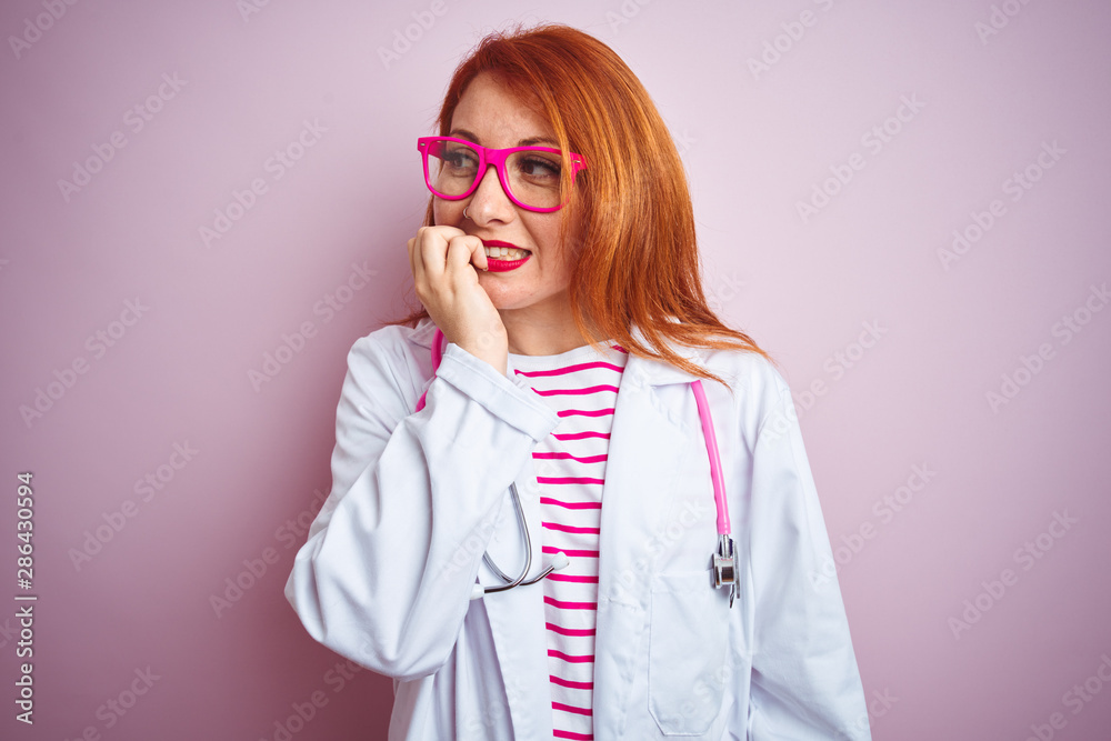 Young redhead doctor woman using stethoscope standing over isolated pink background looking stressed and nervous with hands on mouth biting nails. Anxiety problem.